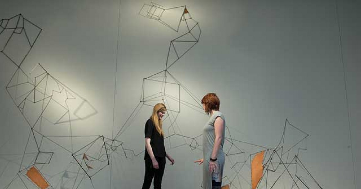 Two students stand in front of a large sculpture made of many segments of straight wire.