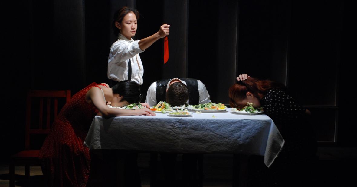  Actor holding red knife over table, while others sit with heads on filled plates