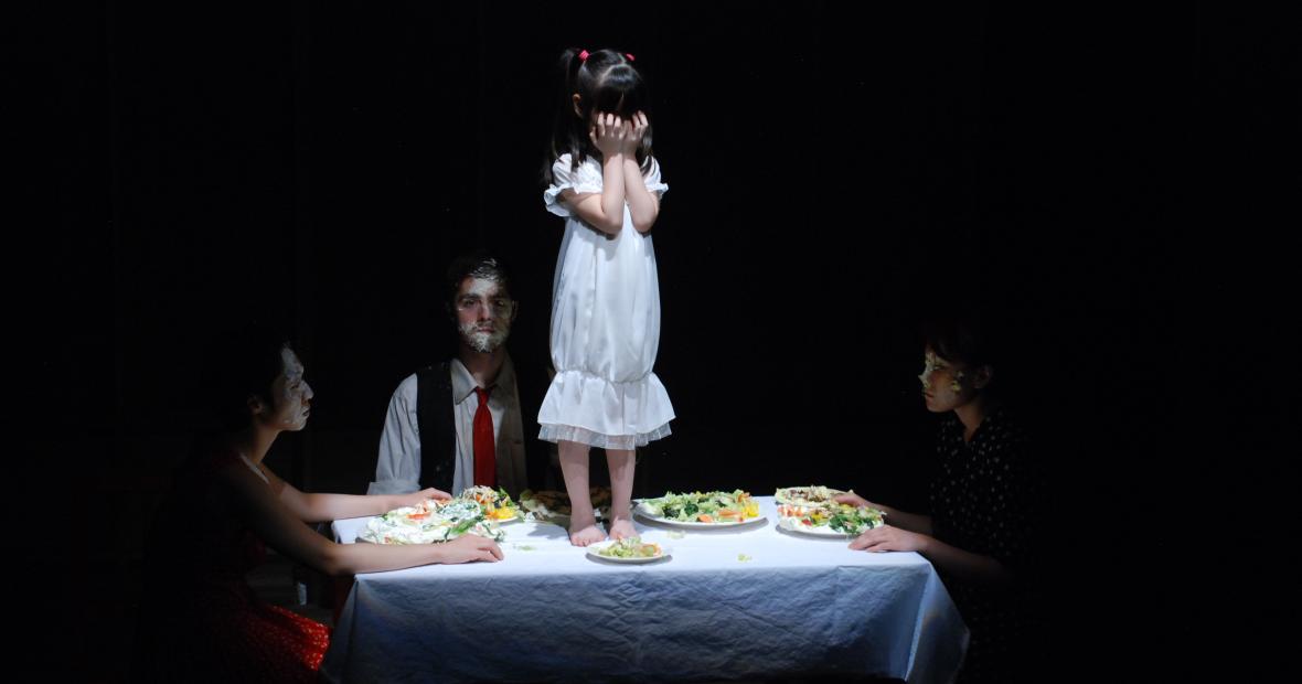 Small girl on table, with other actors seated around her, food on faces