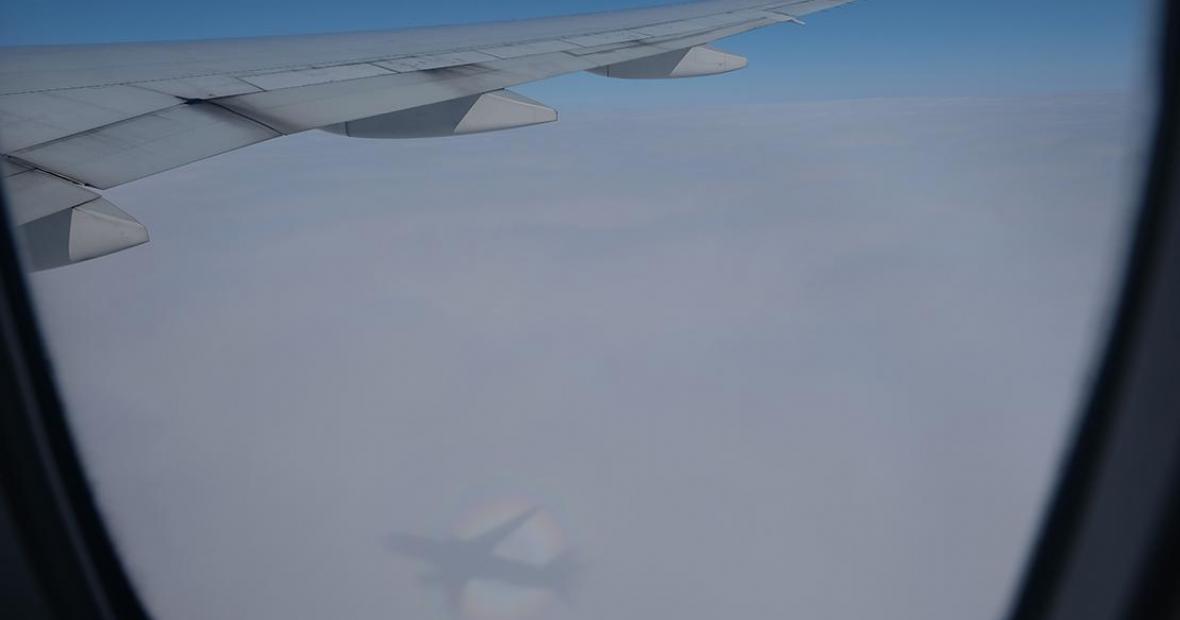 View out of the plane window catches the wing and a shadow of the plane on the clouds below.
