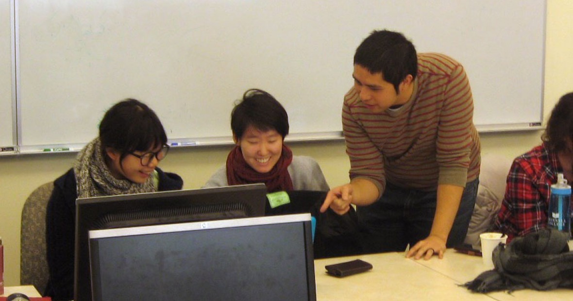 An instructor points to student work on a computer screen while two students look on.