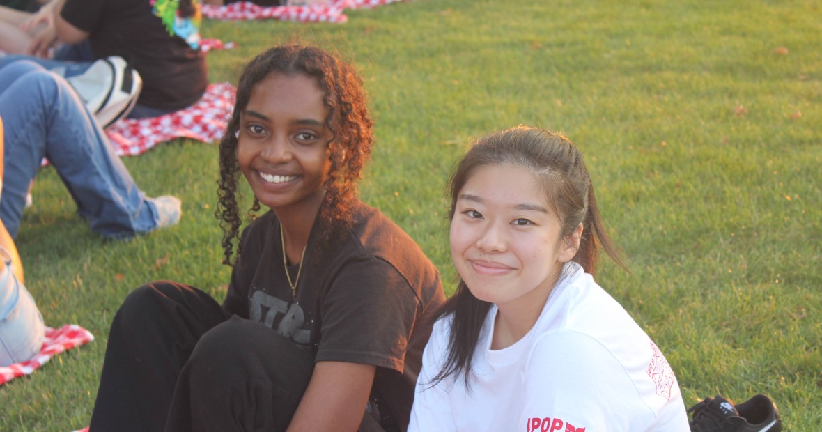 Two students of color sitt on a picnic blanket and smile at the viewer
