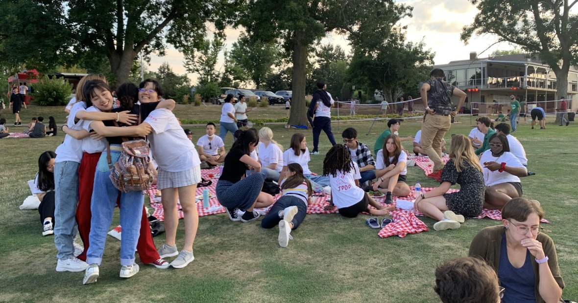 Three students hugging at a picnic while several other people sit and eat on picnic blankets