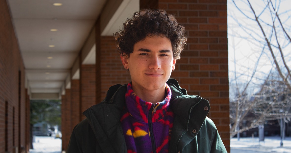A young man with curly brown hair stands outside the Joe Rosenfield Center and smiles slightly. He is wearing a red fleece under a green winter coat.