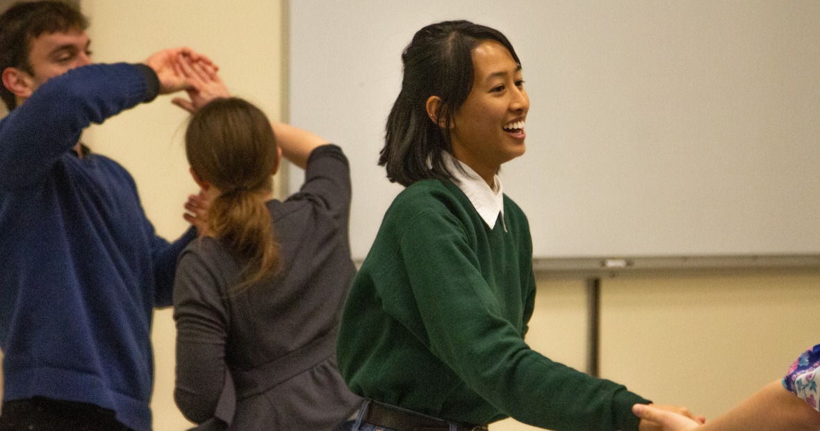 A student in green smiles as they hold an unknown subject's hand. In the background are two students, one in the middle of a turn.