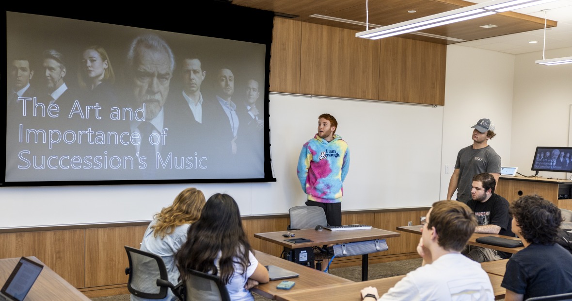A student in a Kenough sweater (pink, blue, and yellow) has his hands behind his back as he looks to his PowerPoint slide. His slide says, "The Art and Importance of Succession‘s music.”
