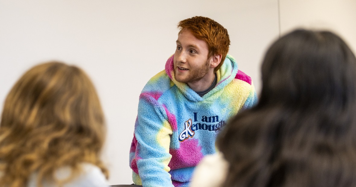 A student in a Kenough sweater (pink, blue, and yellow) leans over a desk to presumably begin his presentation. He looks up towards an off-screen PowerPoint slide and smiles.