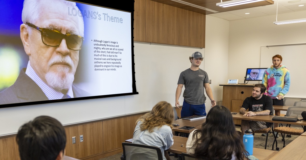 A student in a gray shirt and gray cap points to a PowerPoint screen that reads, "Logan's Theme (Cont'd)," and is followed by two long chunks of text.