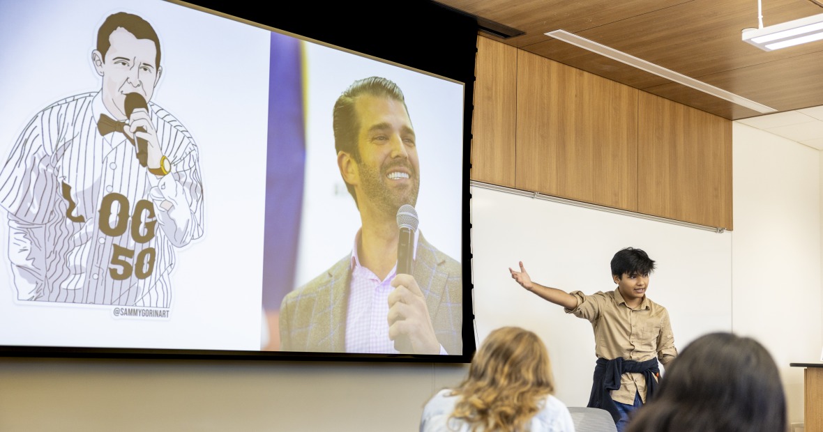 A student with black hair and a brown shirt points to a PowerPoint screen, with a drawn image of Kendall Roy rapping, alongside an image of Donald Trump, Jr.