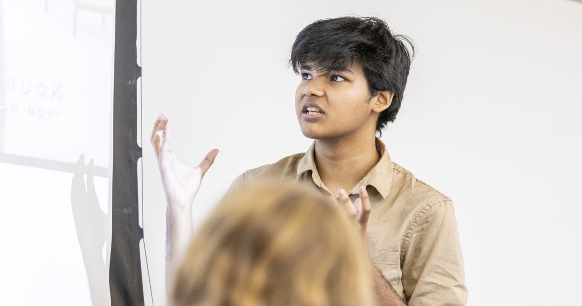 A student in a brown shirt looks at a PowerPoint slide as he gestures.