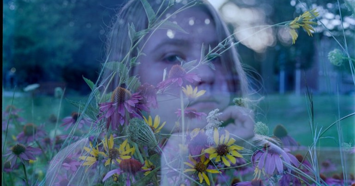 An image of a woman with her hand on her chin. She is transparent and is behind a transparent picture of flowers, grass, and the sky.