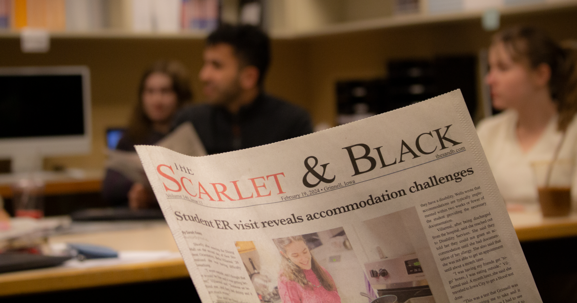 A closeup of the front page of the Scarlet and Black newspaper, while newspaper staff discuss the issue in the background.