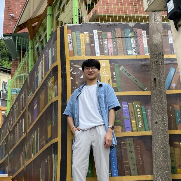 Daniel stands in front of a painted multicolored bookshelf with a hand in his pocket, smiling at the viewer