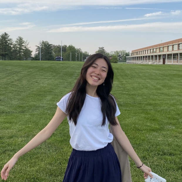 Kana stands in a green field with her arms outstretched, smiling at the viewer