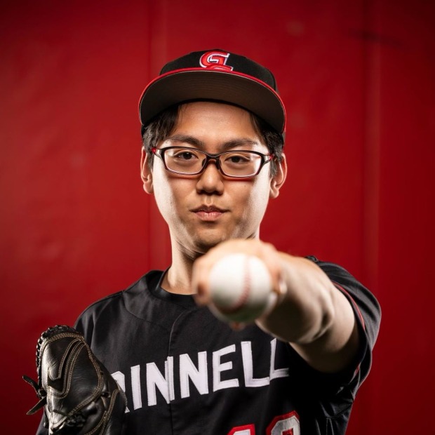 Kenji wears a Grinnell baseball uniform, hat, and baseball glove, and points a baseball straight up towards the viewer