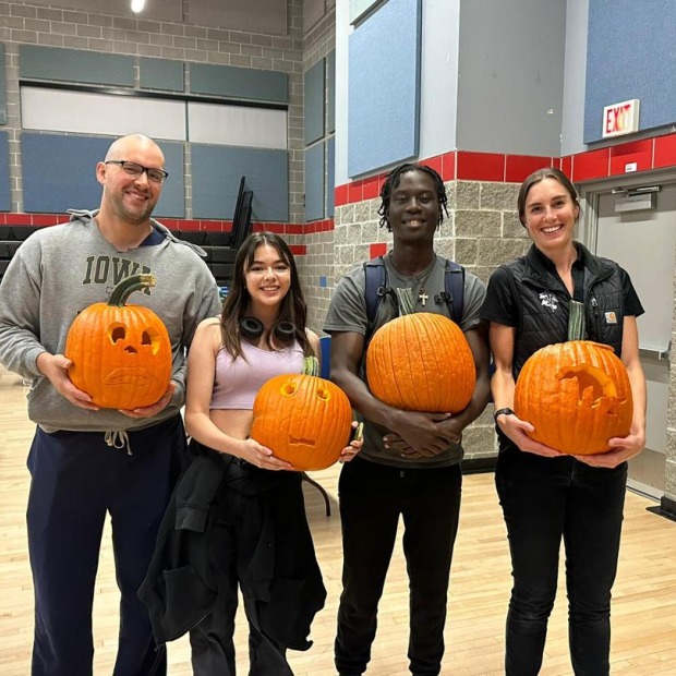 Four individuals hold pumpkins, smiling at the viewer