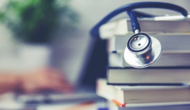 Focused shot of stethoscope draped over books with unfocused hand typing on laptop in background