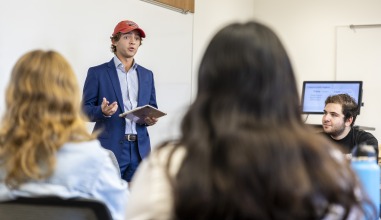 A student in a navy suit and blue dress shirt (and a red cap), holds an IPad as he talks to the class.