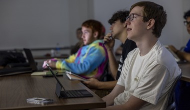 The room is dark. Three students are seated in a line, with their heads tilted up watching a screen, whose light shines on their faces.