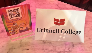 On a marble table lies a name tag that reads, "Grinnell College," with the Grinnell College logo. Next to the tag is a Hamburger Mary's sign with a QR code.