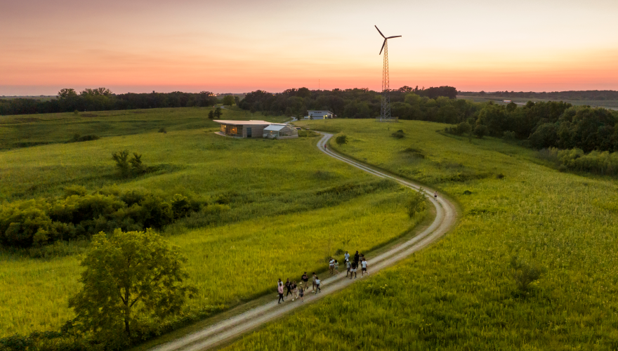 Aerial view of green fields at sunset. a group walks down a winding road towards buildings and a wind turbine