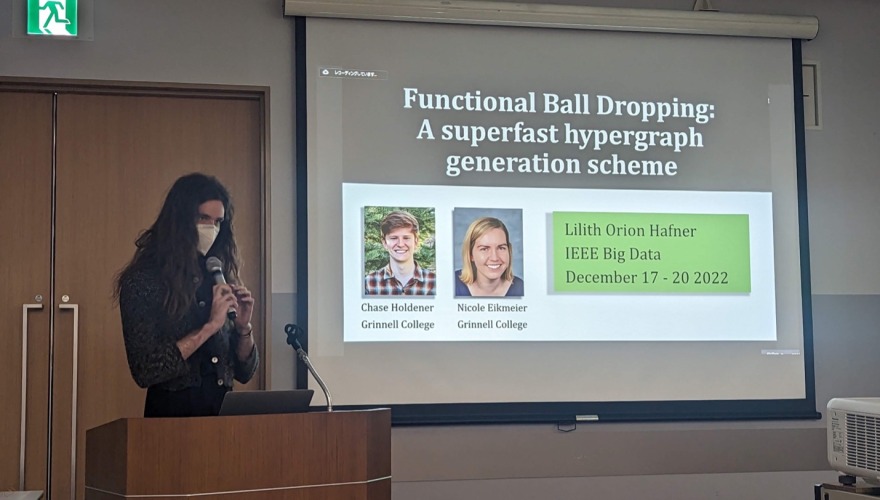 A student with long brown hair stands behind a podium with a microphone. They gesture to the screen, where the words "Functional Ball Dropping: A superfast hypergraph generation scheme" are projected.