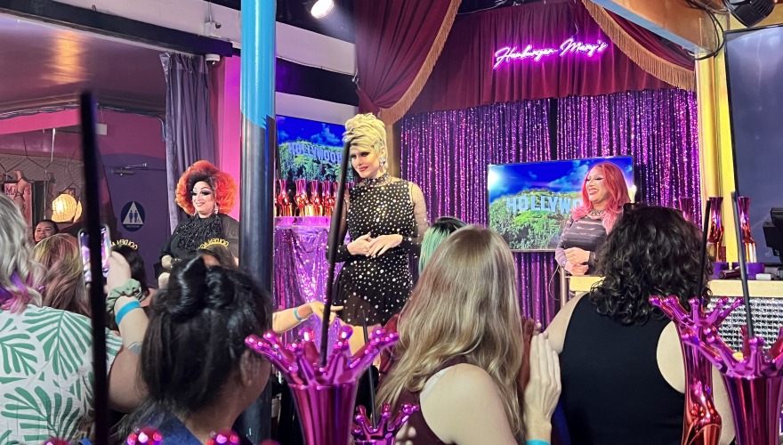 Three drag queens stand in front of TV screens, a purple sequined background, and Hamburger Mary's sign. 