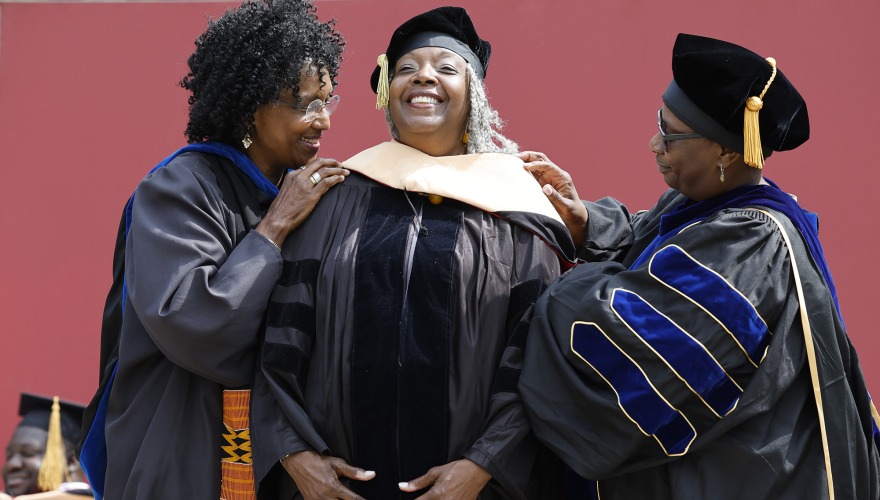 A woman smiles broadly as two colleagues place an academic stole over her head in honor of her honorary degree.