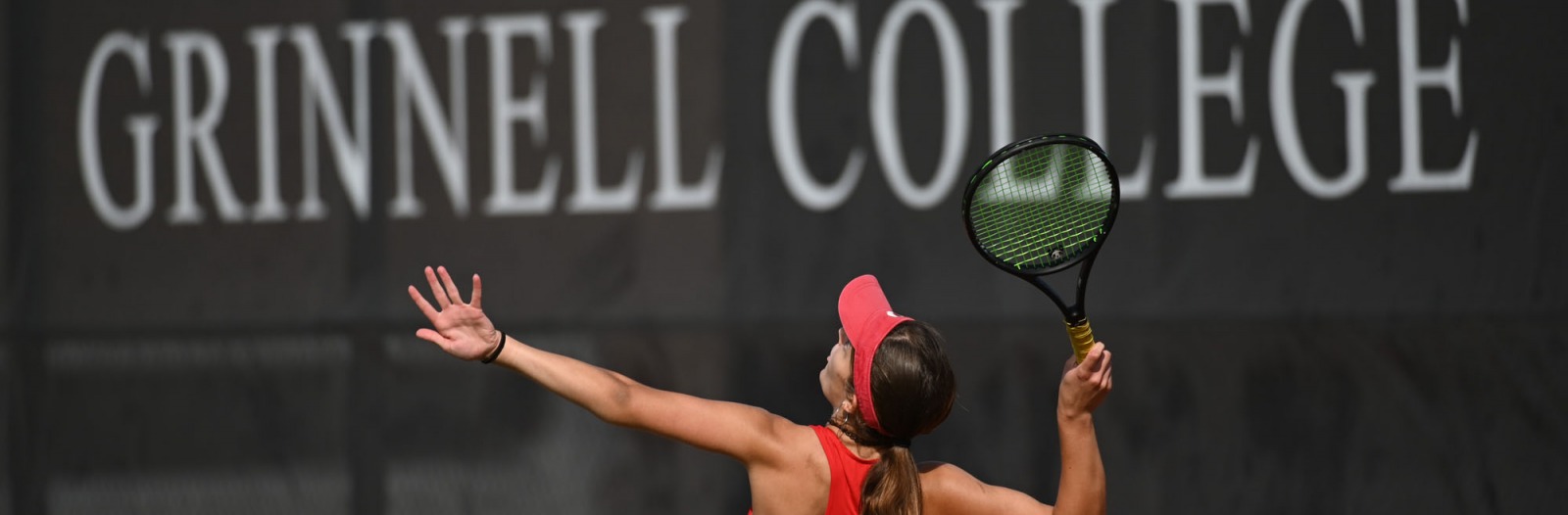 tennis player serves in front of ‘Grinnell College’ sign