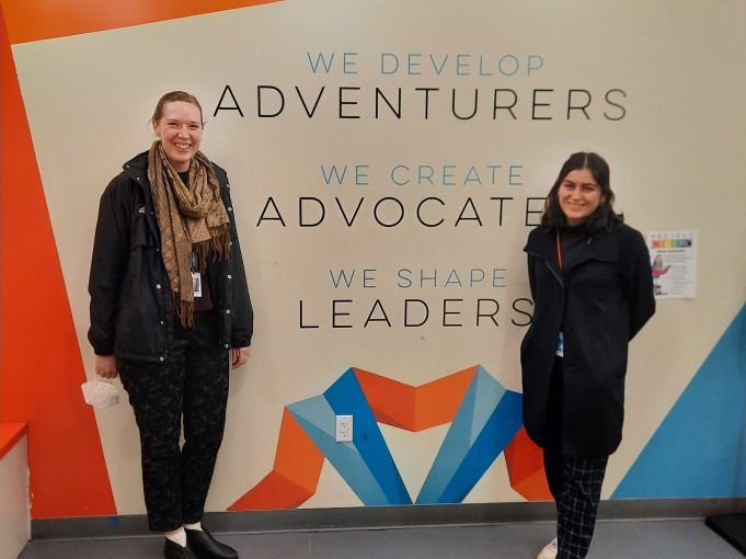 Vanessa and her extern host stand in front of a branded orange and white wall that says "We develop adventures, We create advocates, We shape leaders."