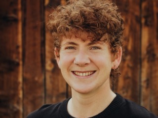 Crys, a young person with curly short hair, wears a black shirt and smiles in front of a wooden background. 