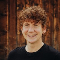 Crys, a young person with curly short hair, wears a black shirt and smiles in front of a wooden background. 