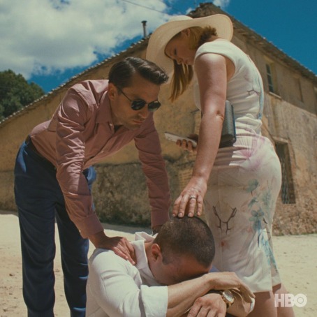 A screenshot from Succession: Roman, in a pink dress shirt, touches Kendall's shoulder, who is seated on the ground with his head in his arms. Shiv, with her back turned to Kendall, turns her head towards him and touches his head.