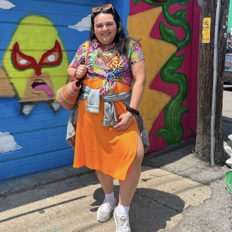 A Latina woman in a colorful neon shirt and orange skirt poses in front of a mural on a garage door. 