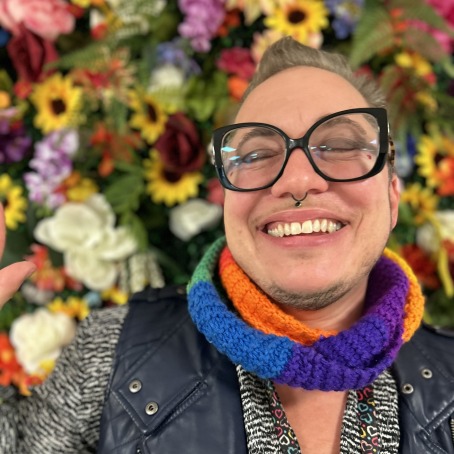 A white person with a nose ring and bold black glasses smiles at the camera. Xe stands in front of a flower background.