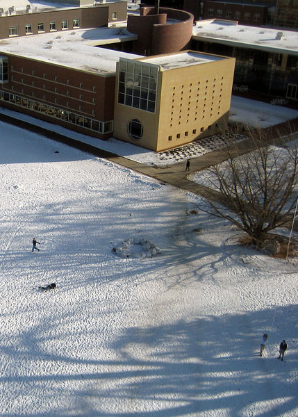 View of snowy Mac field, with Rosenfield Center seen from above near the top of the photo. Figures are playing and flying the kite. Kite string is visible on the right.