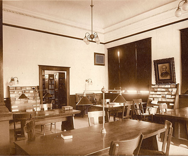 Internior of Carnegie periodical reading room, shows large room with lots of tables, lamps, and periodical shelving