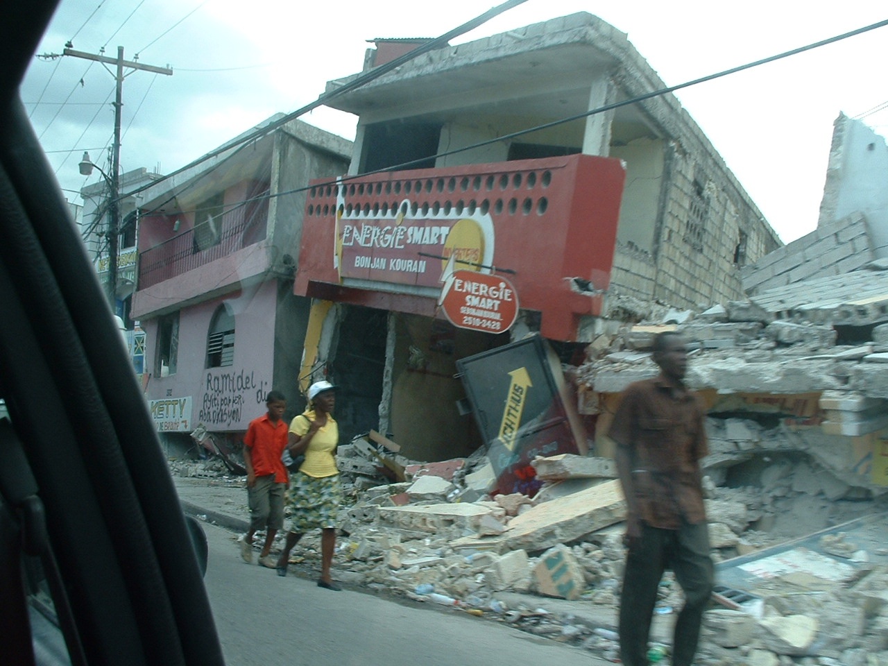 Three people walk down a street with ruined, collapsed buildings in the background