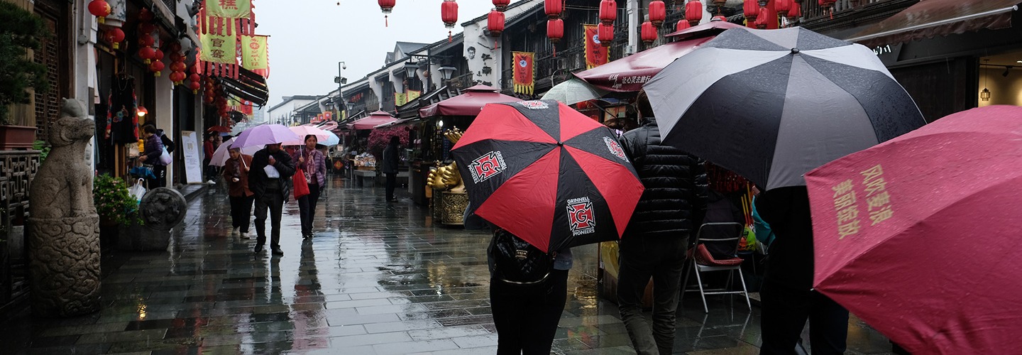 Red lanterns are suspended over a street with many pedestrians carrying umbrellas. One is black and red and displays the honor G