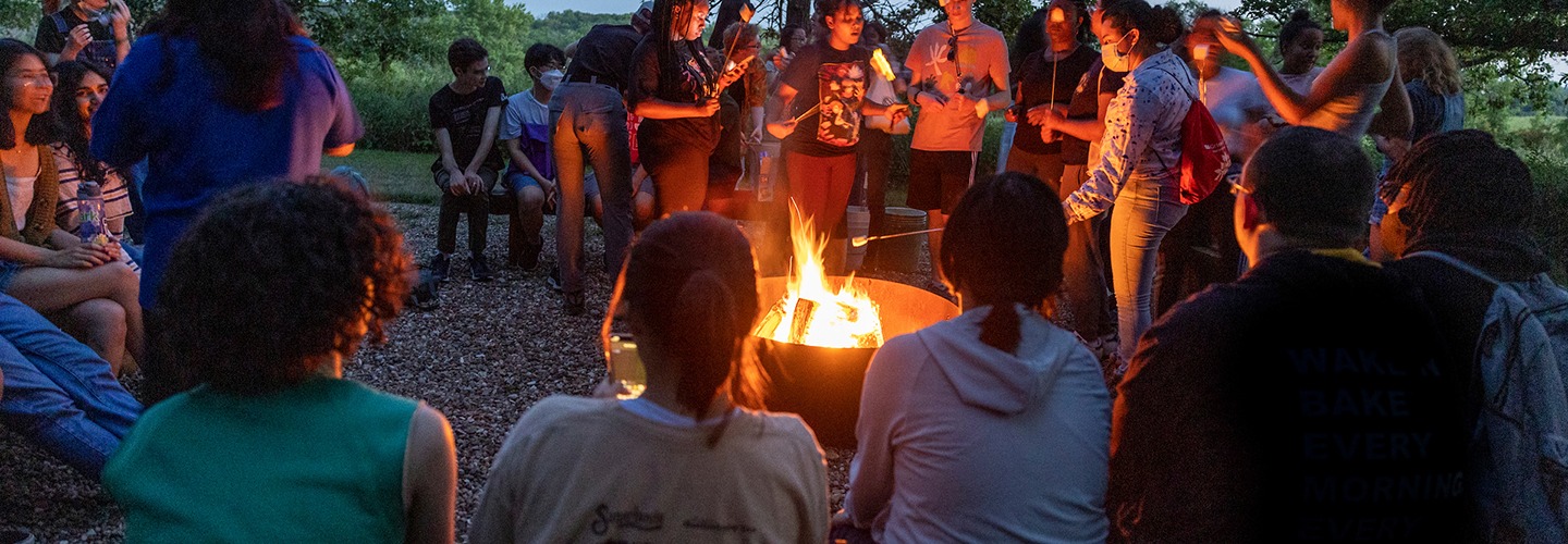 A group of students gathers in a ring around a glowing bonfire in a wooded clearing at dusk. Some sit on benches in the foreground while others stand, roasting marshmallows on long sticks. The warm firelight illuminates their faces against the darkening evening sky and surrounding trees.