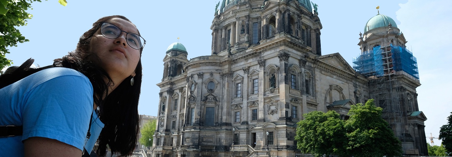 A student wearing glasses and a blue shirt leans over a stone balustrade on a sunny day, gazing across a river toward the grand, green-domed Berlin Cathedral. A white tour boat glides through the water, with lush green trees lining the riverbanks.