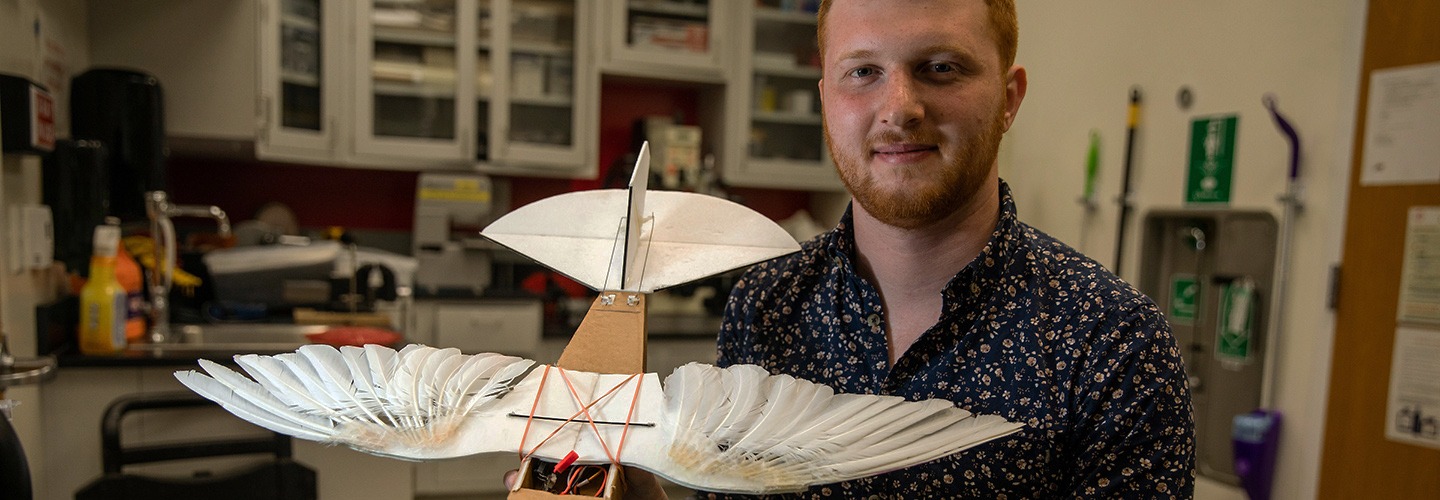A student with short red hair and a beard smiles at the camera inside a lab, holding a custom-built mechanical bird model. The device features realistic white feathered wings stretched wide, bound to a brown chassis with orange bands above a workshop table.