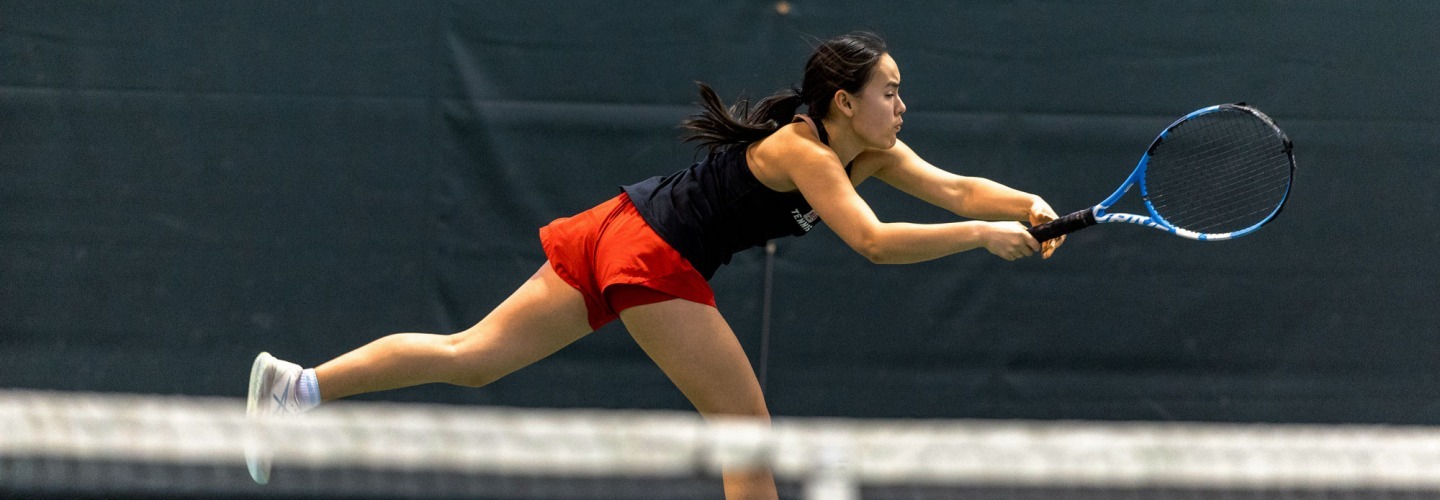 An athlete in a black sleeveless top and red shorts lunges forward in a low stance across a tennis court to make a two-handed backhand stroke. The white net stretches across the immediate foreground against a dark background wall.