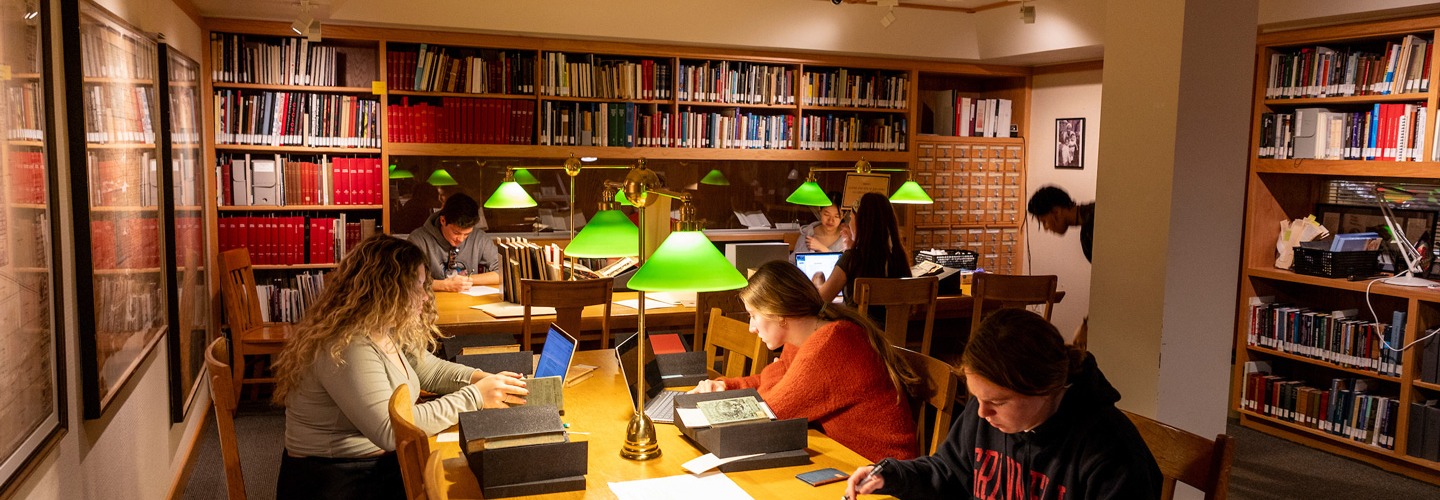 Students sit at a long wooden table in a warmly lit archives room, studying old books and working on laptops. Green-shaded desk lamps cast bright light over their workspaces, surrounded by wall-to-wall bookshelves filled with volumes and framed artwork.