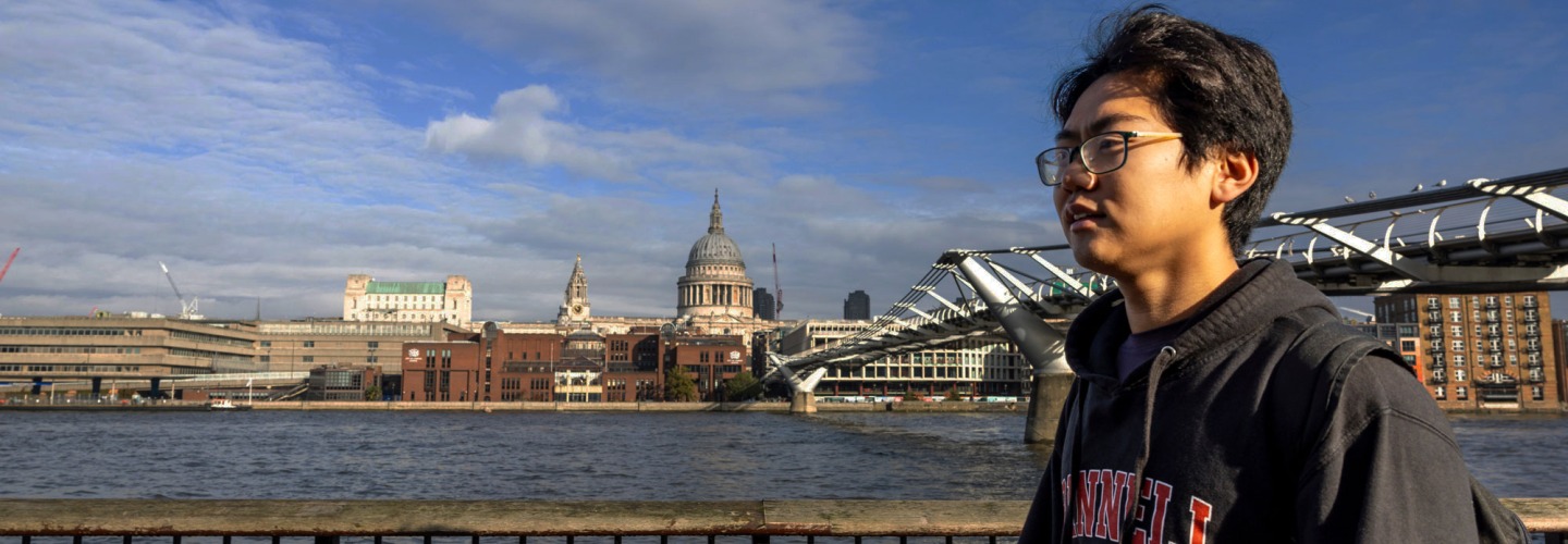 A student in a Grinnell College sweatshirt stands at the side of a river. A bridge to a city in the distance is visible behind him.