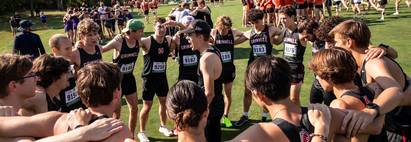 The Grinnell men's cross country team, in uniform, huddles before a competition. Other teams are visible on the field behind them.