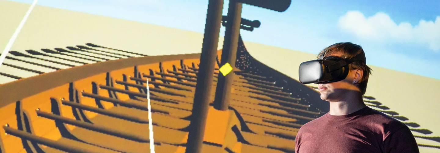 Man in VR headset with controls in hand stands in front of screen showing the longship he is exploring