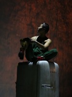 A performer in overalls perches atop an aged vintage refrigerator under dramatic light