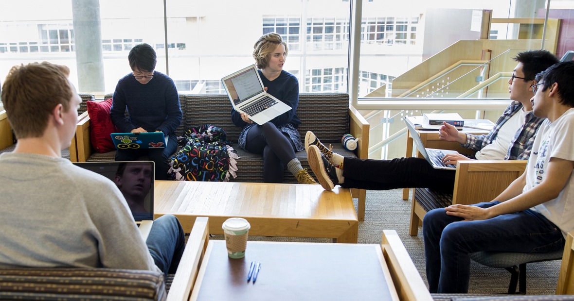 A group of students sit and type while one shows her laptop screen to another.