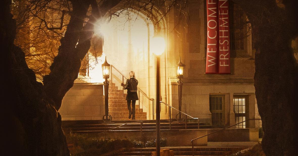 Young woman walks up stairs under lights on dimly lit campus as &quot;Welcome Freshman&quot; banners hang nearby. 